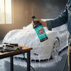 Planet Car Care Foam Fury snow foam shampoo bottle held by a person in a garage with a foam-covered car in background